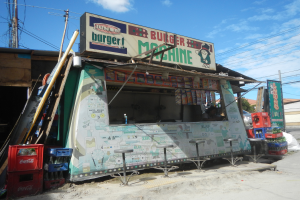 Ein Foodtruck steht am Rande einer Stra├če mit einer Person darin, umgeben von Hockern, Kisten und anderen Gegenst├Ąnden, und einer Tafel mit Text, mit Strommasten, Geb├Ąuden, B├Ąumen und einem bew├Âlktem Himmel im Hintergrund.