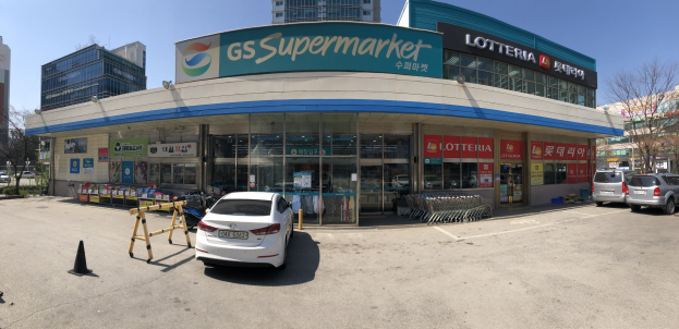 Ein Auto vor einem "GS Supermarkt"-Geschäft mit anderen Fahrzeugen, Verkehrskegeln und Gegenständen auf der Straße, Gebäuden, Bäumen und einem klaren blauen Himmel im Hintergrund.