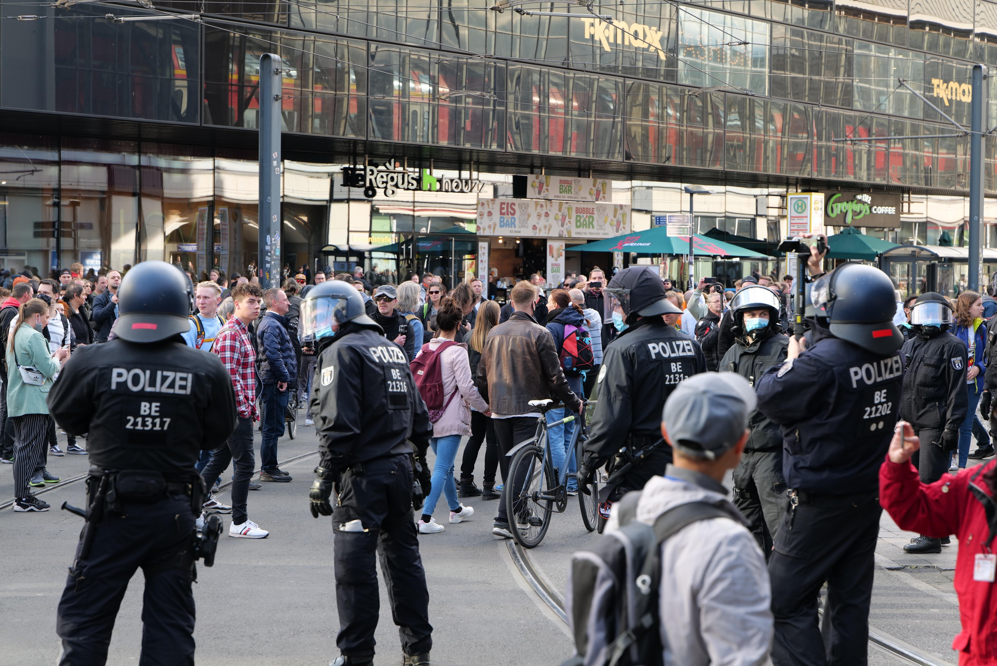 Eine Gruppe von Polizisten vor einer Menschenmenge auf der Straße, wobei einige Personen Helme tragen und Taschen oder Fahrräder haben, und im Hintergrund Gebäude mit Glaswänden und Schildern.