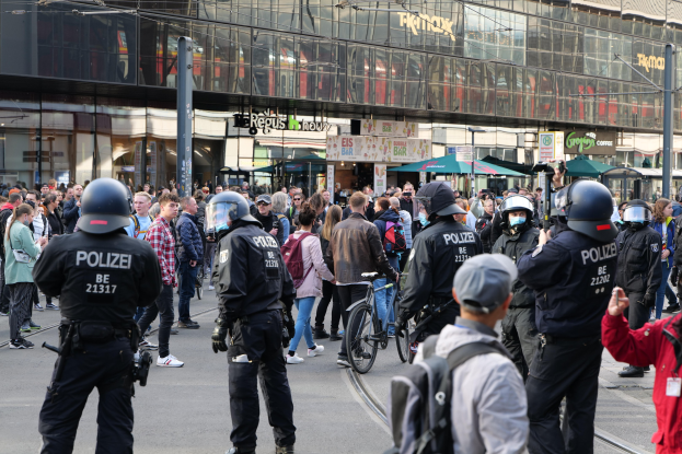 Eine Gruppe von Polizisten vor einer Menschenmenge auf der Straße, wobei einige Personen Helme tragen und Taschen oder Fahrräder haben, und im Hintergrund Gebäude mit Glaswänden und Schildern.