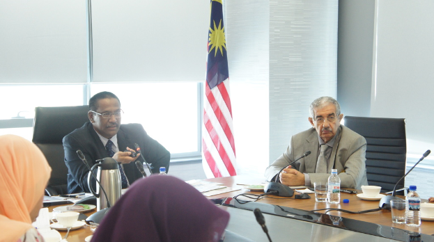 Eine Gruppe von Menschen, darunter Männer in Anzügen und zwei Frauen in Burkas, sitzt um einen Tisch mit Wasserflaschen, Mikrofon, Tassen und einem Teekessel; im Hintergrund ist ein Fenster mit Vorhängen und einer Flagge zu sehen.