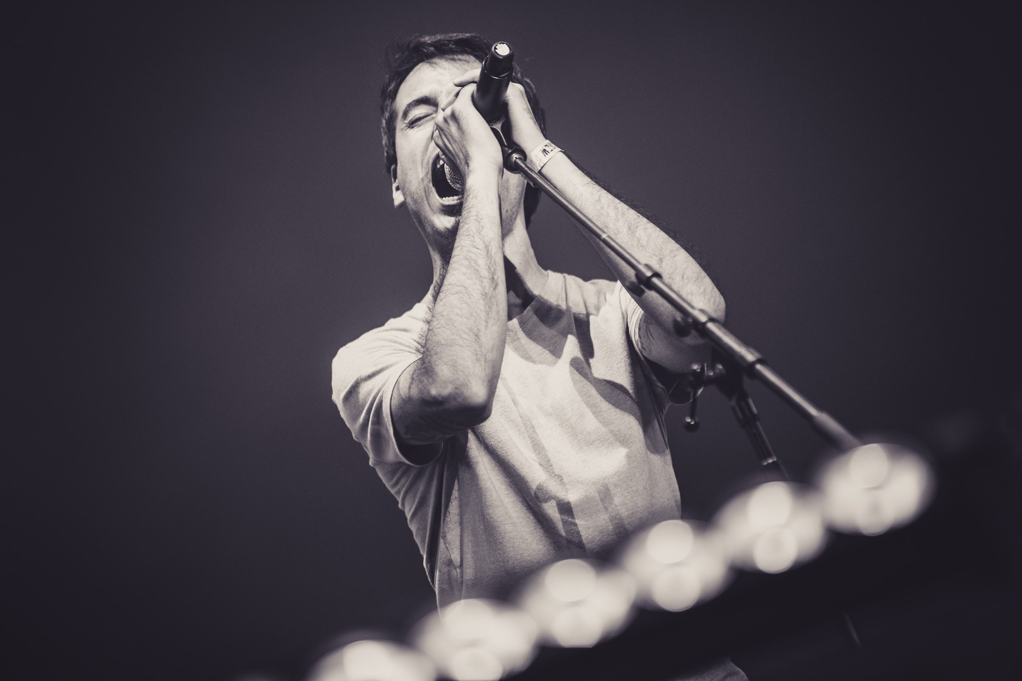 Ein Mann, der in ein Mikrofon singt, auf einer hell erleuchteten Bühne, trägt ein schwarzes T-Shirt und Jeans, hat eine unordentliche Frisur und hat die Augen geschlossen.
