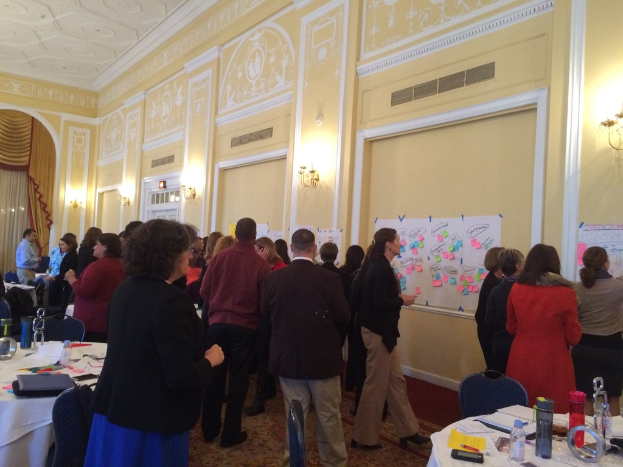 Eine Gruppe von Menschen untersucht Plakate und Charts, die an einer Wand in einem Raum aufgehängt zu sein scheinen, der wie ein Kunstmuseum aussieht.