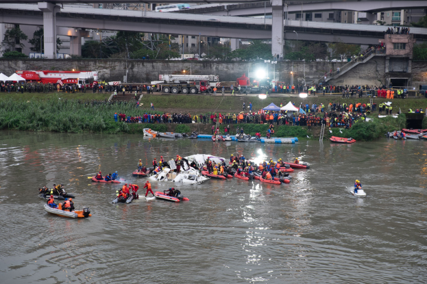 Eine Gruppe von Menschen in kleinen Booten, die Lebenswesten tragen, treiben auf einem Gewässer, umgeben von Pflanzen, Gras, Zelten, Fahrzeugen, Laternenmasten, Bäumen, Gebäuden und einer Brücke im Hintergrund, was auf einen Rettungseinsatz hindeutet.