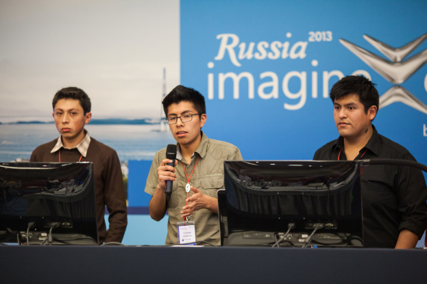 Drei Jungen stehen neben einem Schreibtisch mit zwei Computersystemen, einer spricht in ein Mikrofon und trägt ID-Karten mit roten Tags, vor einer Wand mit einer Werbetafel, auf der 'Russia Imagine 2013' steht.