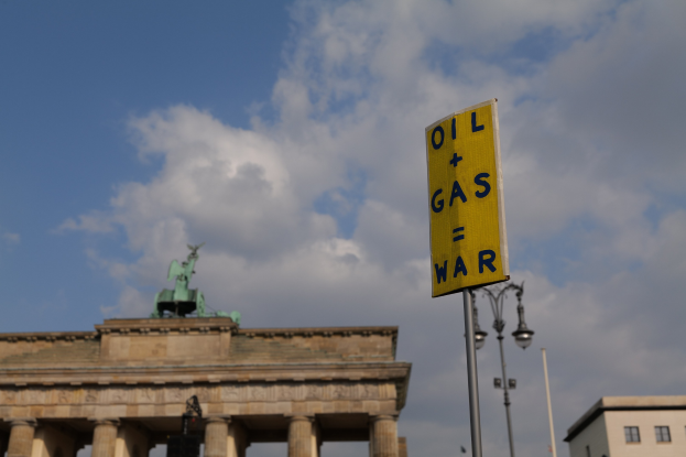 Gelbes Schild mit der Aufschrift "Öl- und Gaskrieg" vorne mit dem Brandenburger Tor und Berliner Gebäuden im Hintergrund.