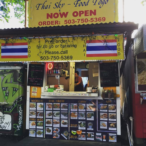 Ein Geschäft mit verschiedenen Artikeln wie Getränkedosen, Speisekarten, einer Uhr, Gewürzen in Plastikbehältern, Gewürzen, Werbetafeln, Namensschildern und einem Blick auf den Himmel.