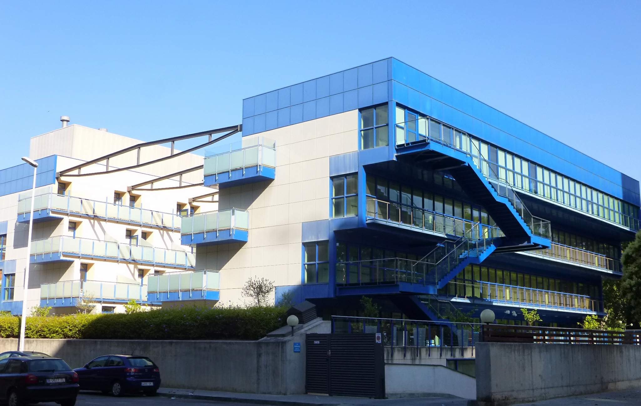 Großes modernes Gebäude mit zahlreichen parkenden Autos davor, umgeben von Straßenlaternen, Gittern, Grünflächen und einem klaren blauen Himmel, als neuer Hauptsitz der University of California, Berkeley bezeichnet.