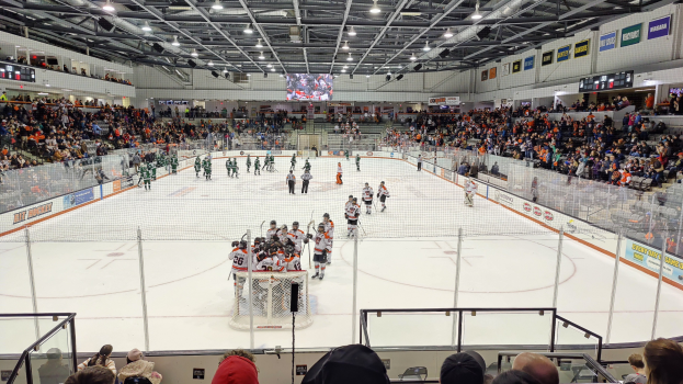 Eishockeyspiel in einer großen Halle mit Spielern auf dem Eis, einem Tor, Zuschauern in den Rängen, Deckenbeleuchtung und Bildschirm sowie Bannern darüber.