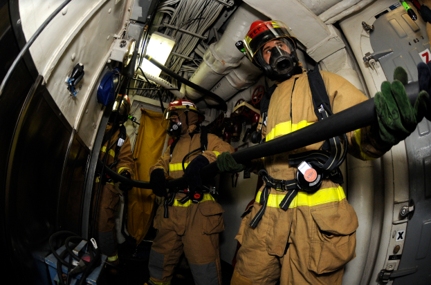 Eine Gruppe von Feuerwehrleuten in Helmen und Handschuhen hält einen Schlauch am Heck eines Flugzeugs, mit Kabeln, Lampen und anderen Gegenständen im Hintergrund und Text am unteren Bildrand.