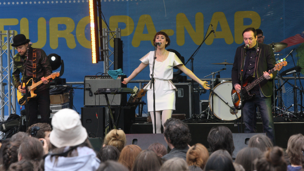 Eine Bühne mit vier Musikern: Eine Frau singt in einem weißen Kleid in der Mitte, zwei Männer spielen Gitarren auf der linken und rechten Seite und ein Publikum und Lautsprecher im Hintergrund.