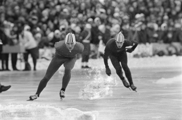 Zwei Männer im Geschwindigkeits-Eisschnelllauf auf einer Eisbahn vor Publikum, beide tragen Helme und Eislaufschuhe, mit unscharfem Hintergrund und einem sichtbaren Bein auf der linken Seite, in Schwarz-Weiß.