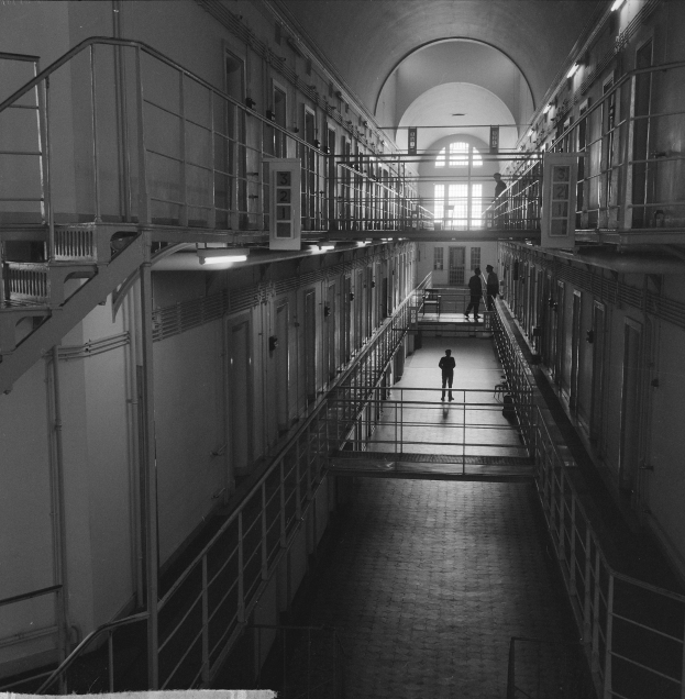 Schwarzes und weißes Foto eines Mannes, der einen langen Gefängnisgang mit Geländern, Türen, Lampen und einem Fenster im Hintergrund entlanggeht.