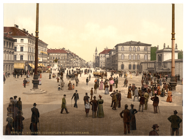 Ein altes Schwarz-Weiß-Foto einer belebten Münchner Stadtstraße aus den frühen 1900er Jahren mit Passanten, einigen mit Schirmen, einem Pferdewagen im Vordergrund, Gebäuden, Bäumen und Pfählen im Hintergrund und Text am unteren Bildrand.