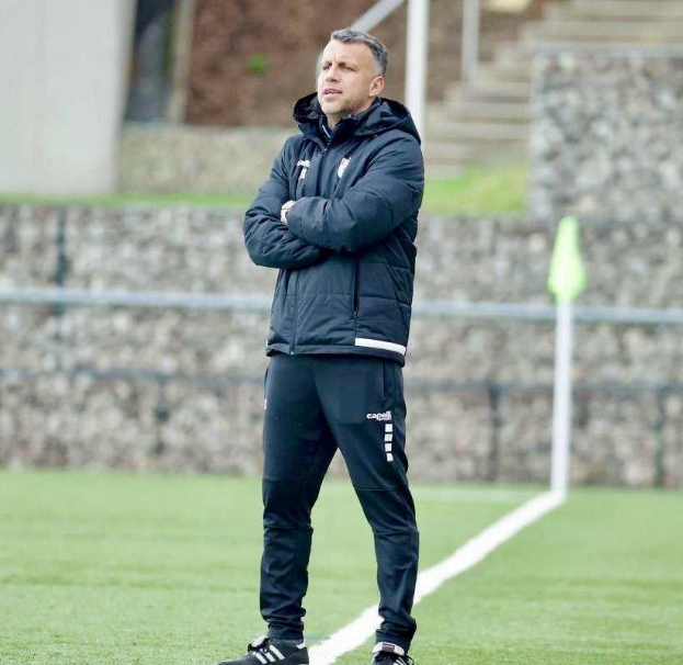 Ein Mann in schwarzer Jacke und Hose steht auf einem Fußballfeld mit verschränkten Armen, der wie ein Trainer während eines Trainings erscheint, mit einem Zaun, Treppe und einer Wand im Hintergrund.