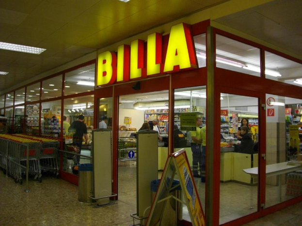 Ein Markt mit einem Eingang in der Mitte, Mülltonnen daneben, einer Reihe von Einkaufswagen auf der linken Seite und einigen Menschen, die im Inneren Produkte anschauen, mit Licht oben.