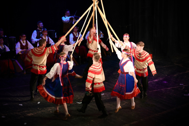 Eine Gruppe von Menschen in bunten Kostümen tanzt um einen Maibaum, einige halten Holzstöcke, mit Musikern im Hintergrund.
