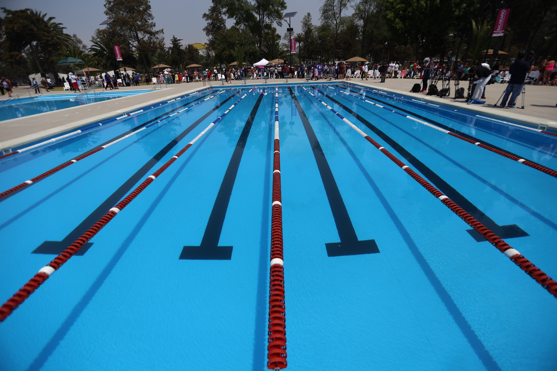 Gro├čes Schwimmbad mit Bahnentrennung, umgeben von Menschen mit Taschen und Gegenst├Ąnden, mit Zelten, Bannern, Masten, einer Anzeigetafel und einem bew├Âlktem Himmel im Hintergrund.