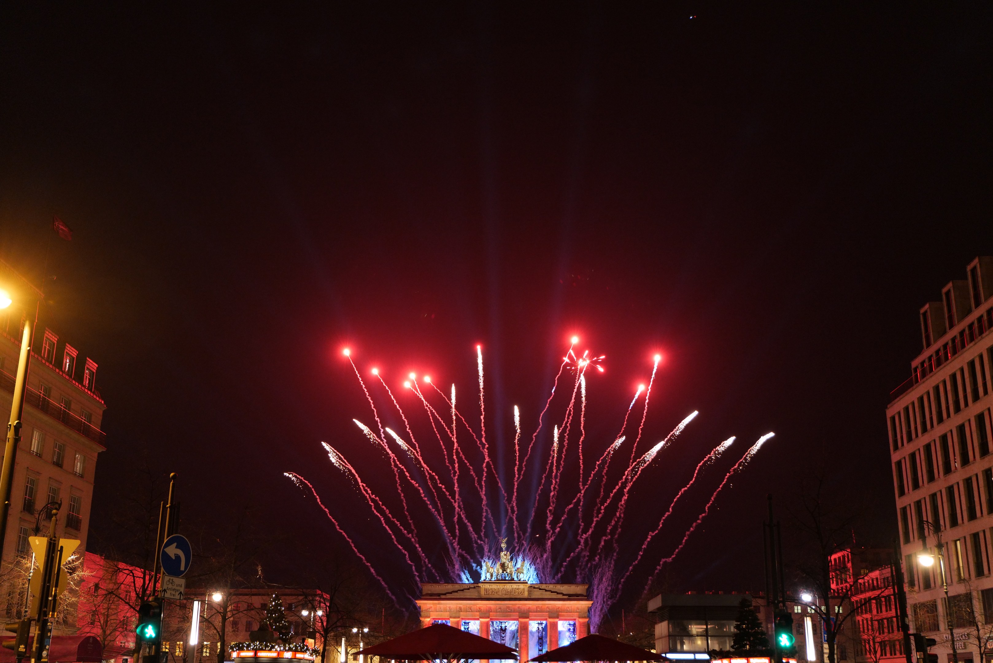 Eine nächtliche Stadtstraße mit dem beleuchteten Brandenburger Tor in Berlin, Deutschland, umgeben von Gebäuden, Bäumen, Laternen, Verkehrsampeln, Schildern, Zelten und Menschen, mit Feuerwerk, das den Himmel erhellt.