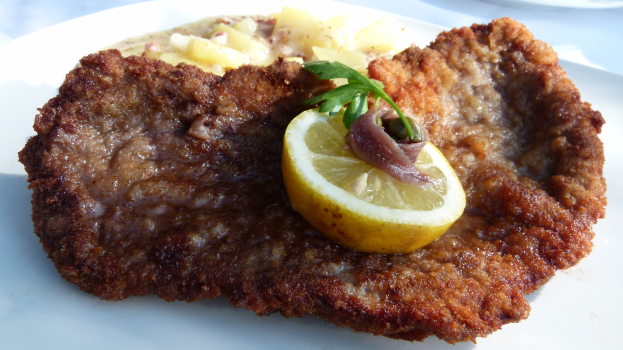 Ein weißer Teller mit einem panierten Schweineschnitzel, Kartoffeln und einer Zitronenscheibe auf einem Tisch.