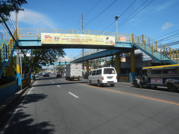 Eine Stadtstraße mit Fahrzeugen unter einer Brücke mit Geländern, Treppen und einem Banner, umgeben von Laternen, Strommasten mit Drähten, Bäumen, Gebäuden und einem bewölkten Himmel.