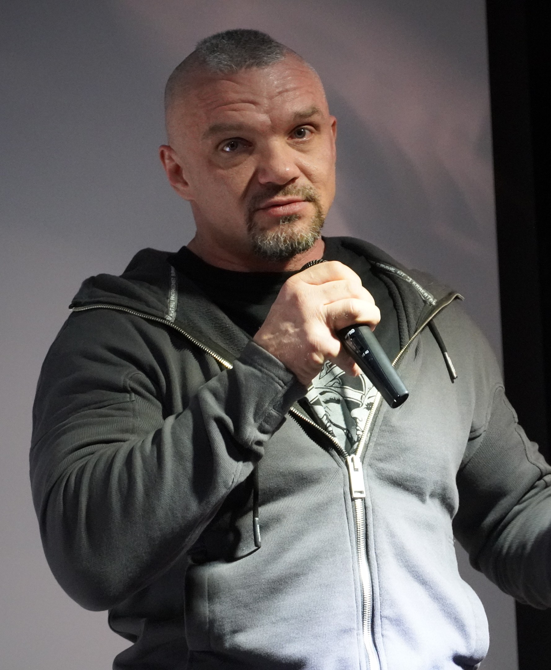 Comedian in einem schwarzen Anzug und wei├čem Hemd, mit Mikrofon haltend, mit zurückgek├Ąmmtem Haar und einem selbstbewussten Ausdruck, vor einer Wand stehend.