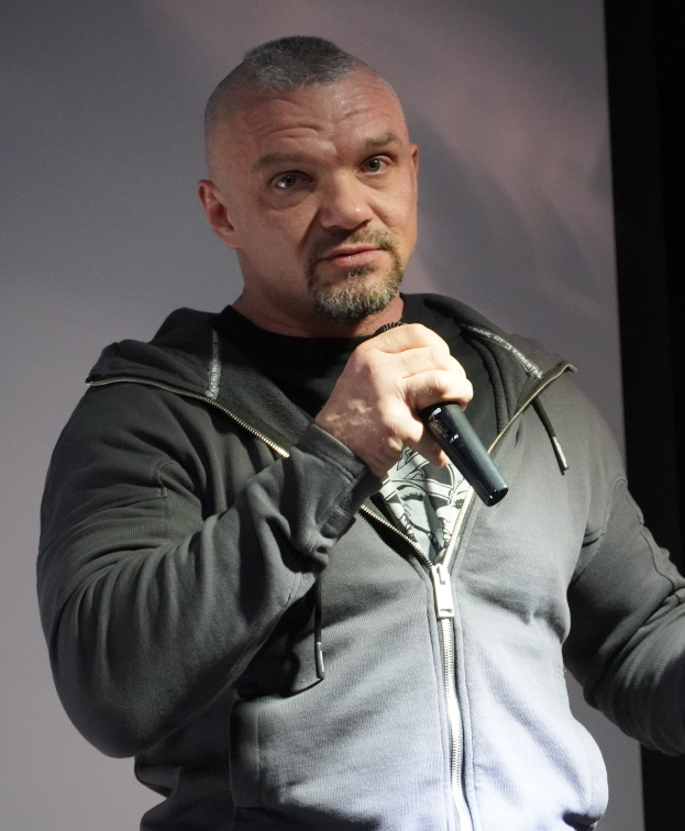 Comedian in einem schwarzen Anzug und wei├čem Hemd, mit Mikrofon haltend, mit zurückgek├Ąmmtem Haar und einem selbstbewussten Ausdruck, vor einer Wand stehend.