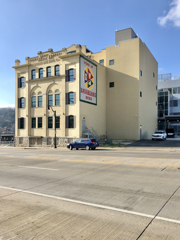 Großes Ziegelsteingebäude mit "Home of Bavarian Brewing Co." -Schriftzug, Straßenmasten, Fahrzeugen, Bäumen, Hügeln und bewölktem Himmel im Hintergrund.