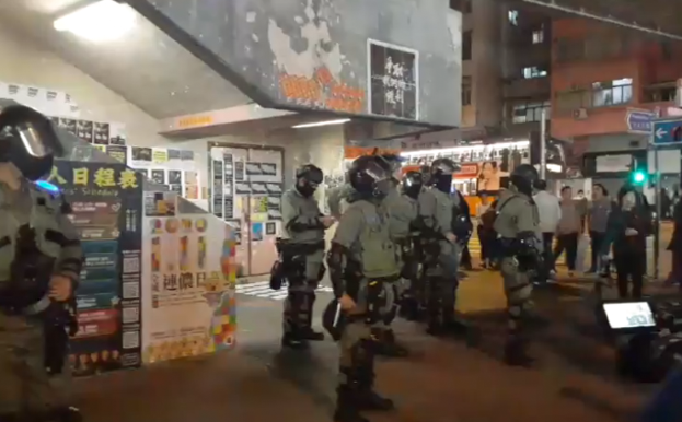 Eine Gruppe von Polizisten in Uniform steht vor einer Menge von Demonstranten mit Helmen und Waffen, mit Plakaten an den Wänden, Lampen an der Decke und Gebäuden mit Fenstern im Hintergrund, in Hongkong.