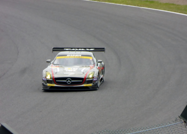 Ein Mercedes-Benz SLS AMG GT3 Rennauto auf einer Rennstrecke mit Text auf dem Auto, einem Maschendrahtzaun unten und grünem Gras im Hintergrund.