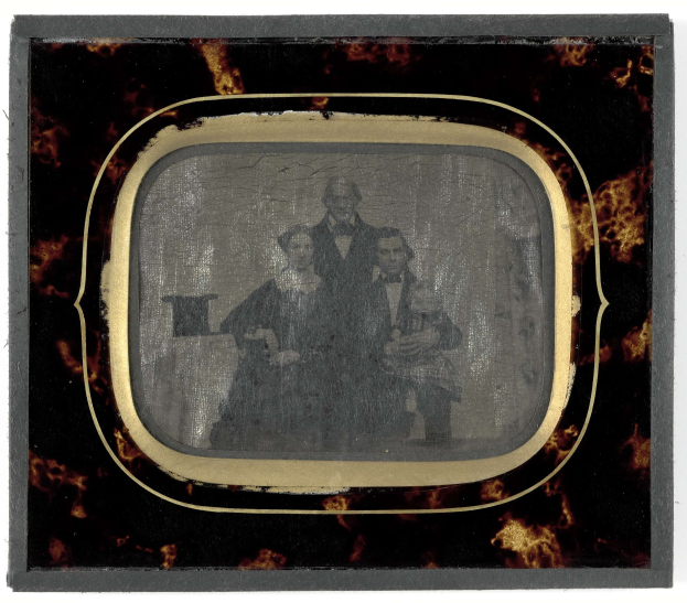 Schwarzes-Weiß-Foto eines Mannes im Anzug und einer Frau in einem unbekannten Outfit, die vor einem alten Fernseher stehen und ernst schauen.