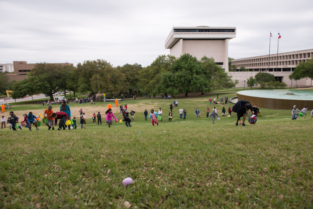 Gruppe von Menschen, die mit Ostereiern in einem grünen Feld mit Bäumen, Gebäuden, Fahnen und einem Brunnen im Hintergrund unter einem bewölkten Himmel spielen.
