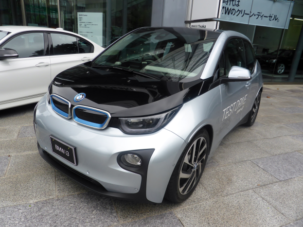 Ein BMW i3 Elektroauto vor einem Gebäude in Tokyo, Japan, umgeben von Glaswänden mit einem Textschild im Hintergrund.