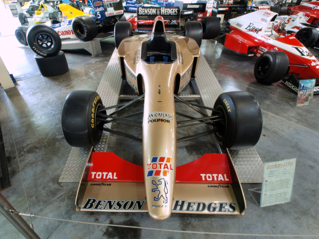 Eine Gruppe von Rennwagen in einem Motorsportmuseum, getrennt durch einen Metallzaun mit einer Texttafel links und Menschen und Gegenständen im Hintergrund.