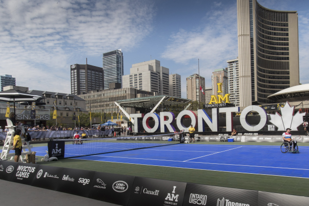 Gruppe von Menschen, die Tennis auf einem Platz mit einer Stadtkulisse im Hintergrund spielen, umgeben von Zuschauern, Bannern, Fahnen und Bäumen unter einem bewölkten Himmel.