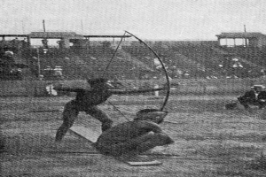 Zwei Männer auf einem Feld, die Bogenschießen üben, einer hält einen Bogen und einen Pfeil in der Hand, mit ein paar Menschen im Hintergrund und dem Himmel im Bild.