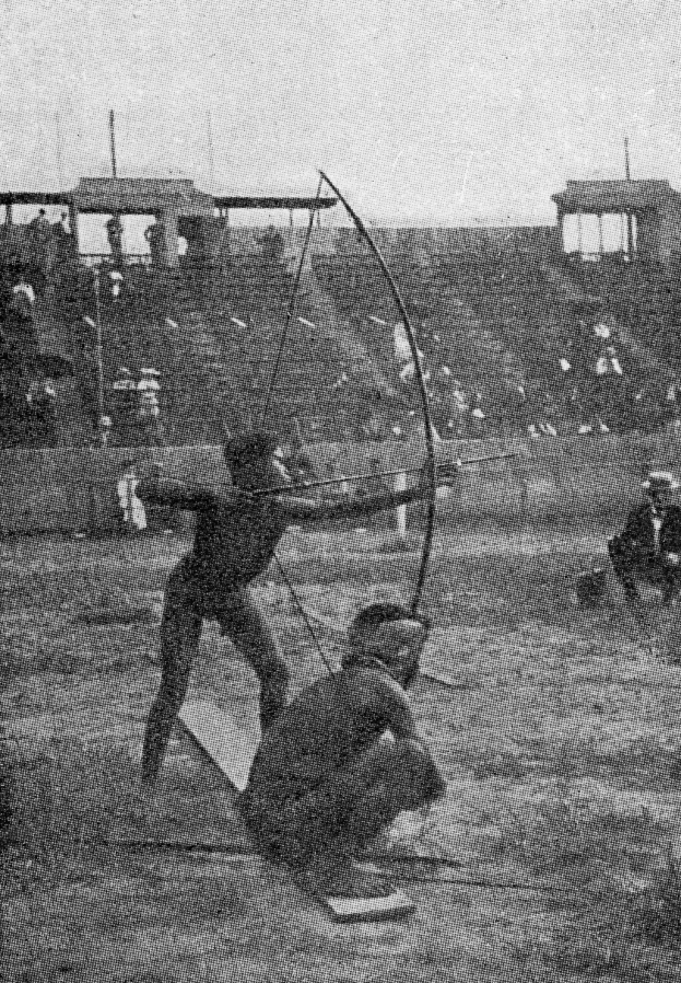 Zwei Männer auf einem Feld, die Bogenschießen üben, einer hält einen Bogen und einen Pfeil in der Hand, mit ein paar Menschen im Hintergrund und dem Himmel im Bild.