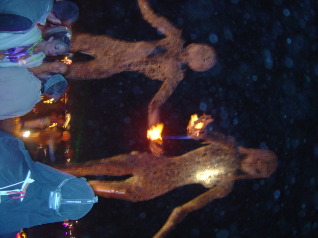 Mehrere Personen stehen auf der linken Seite des Bildes, mit hinter ihnen brennenden Statuen und Lichtern im Hintergrund.