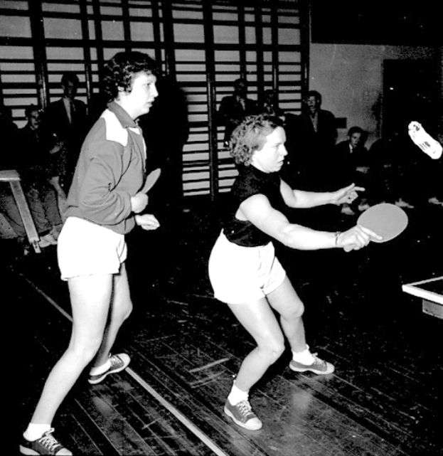 Zwei Frauen spielen Tischtennis auf einem Court, jede hält einen Schläger in der Hand, mit einigen Zuschauern im Hintergrund und einem Tischtennis-Tisch auf der rechten Seite, in Schwarz-Weiß.