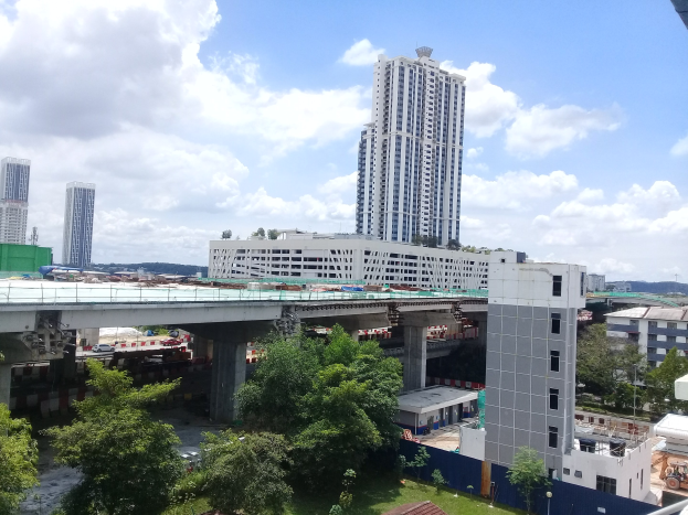 Eine Stadtansicht von der Spitze eines Gebäudes aus, mit Gebäuden, Bäumen, Gras, Fahrzeugen, einer Brücke, Polen und einem bewölkten Himmel, mit einer Baustelle für einen neuen Wohnkomplex im Vordergrund.
