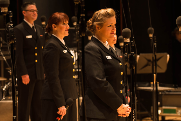 Eine Gruppe von Menschen vor Mikrofonen, mit einem Mann und einer Frau, die Mikrofone halten, und Musikinstrumenten und Ständern im Hintergrund.