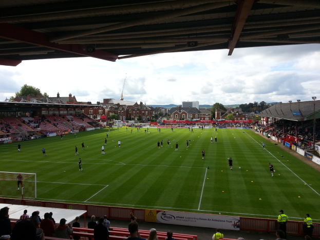 Ein Fußballspiel wird im St Mary's Stadium gespielt, mit Zuschauern auf den Bänken und auf dem Boden stehend, einem Torpfosten links und im Hintergrund Gebäuden, Bäumen und einem bewölkten Himmel.