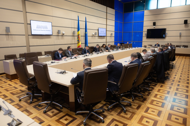 Eine große Gruppe von Menschen um einen langen Tisch mit Mikrofonen und Papieren herum, in einem Raum mit Flaggen, Fernsehern, Lautsprechern und Deckenlampen während einer Sitzung des Europäischen Parlaments in Brüssel.