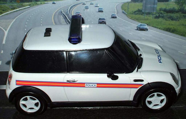 Ein weißer Polizeiwagen ist im Vordergrund zu sehen, hinter ihm bewegen sich mehrere Autos auf der Straße. Rechts und links der Straße sehen wir Gras und Bäume.