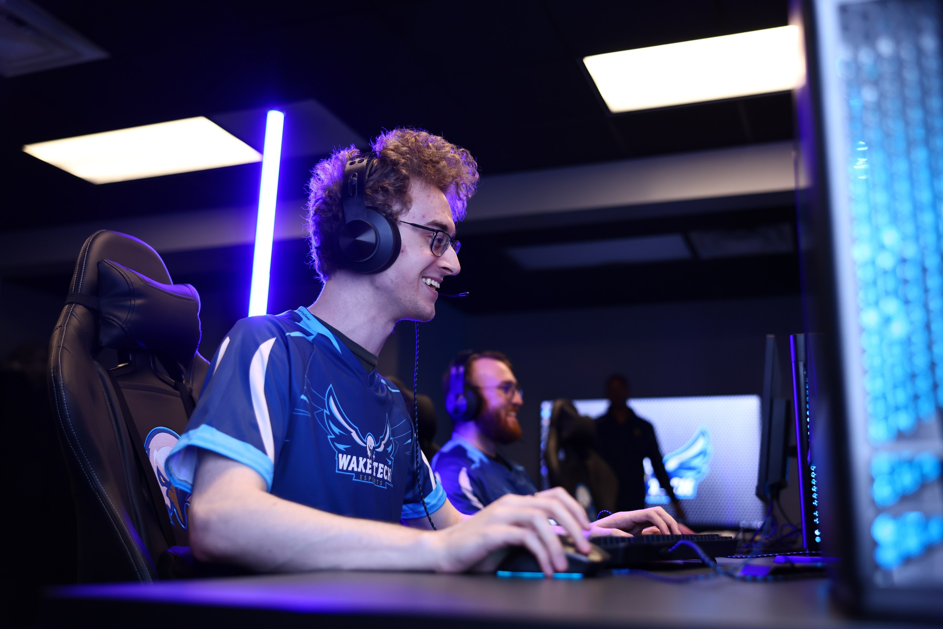 Ein Mann trägt einen Headset und Brille und sitzt an einem Schreibtisch mit einem Computer-Monitor, einer Tastatur und einer Maus und spielt ein Spiel in einem Raum mit einer anderen Person und Deckenleuchten.