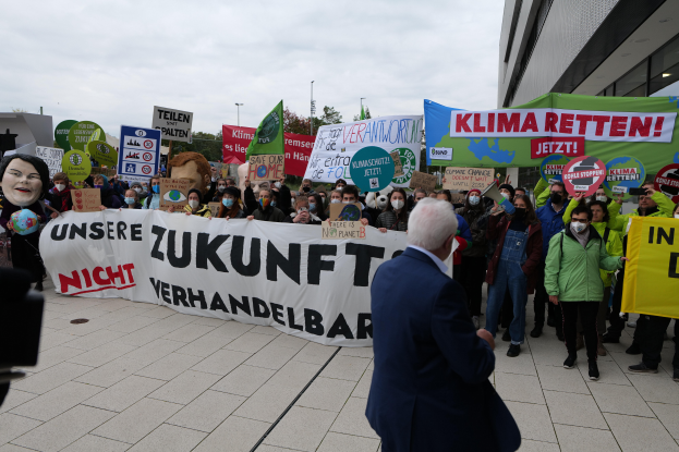 Eine Gruppe von Menschen hält Schilder und Transparente und protestiert gegen den Klimawandel vor einem Gebäude in Deutschland, wobei eine Person mit einer Kamera auf der linken Seite zu sehen ist. Im Hintergrund gibt es Bäume, Pfähle und einen klaren blauen Himmel.