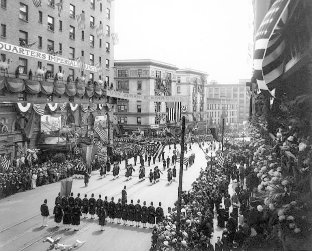 Eine große Menschenmenge marschiert auf einer Straße in New York City während eines Fourth of July-Umzugs, viele tragen Kappen und halten Fahnen, mit Pfählen, Bäumen und Gebäuden mit Fenstern im Hintergrund und lesbarem Text auf einem Gebäude.
