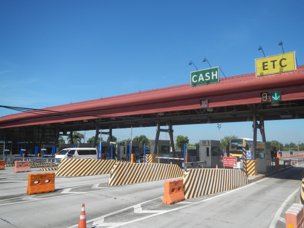 Eine Mautstelle mit einem "Bar"-Schild, umgeben von Fahrzeugen, Schrankenstangen, Hinweistafeln, Straßenlaternen, Strommasten, Stromkabeln, Bäumen und einem bewölkten Himmel.