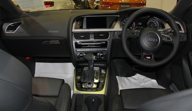 Das Innere eines Audi-Fahrzeugs mit einem Lenkrad, Armaturenbrett mit Tacho, Ganghebel, Sitzen und einer Musikanlage, mit anderen Autos und Menschen im Hintergrund.