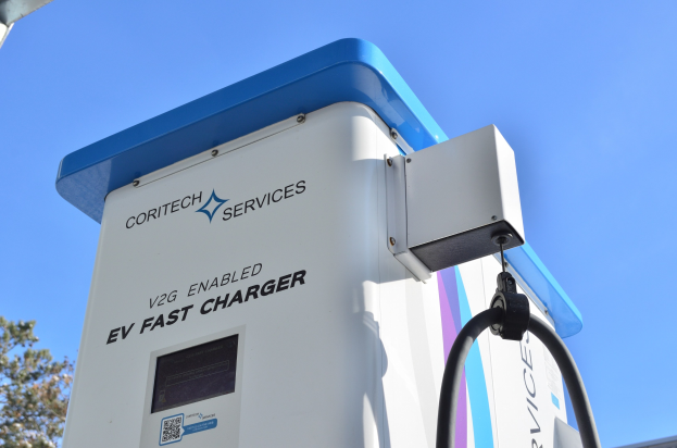 Elektrisches Fahrzeug-Ladestation mit einem blauen und weißen "V2G Fast Charger"-Schild, umgeben von Bäumen und einem Gebäude unter einem klaren blauen Himmel.